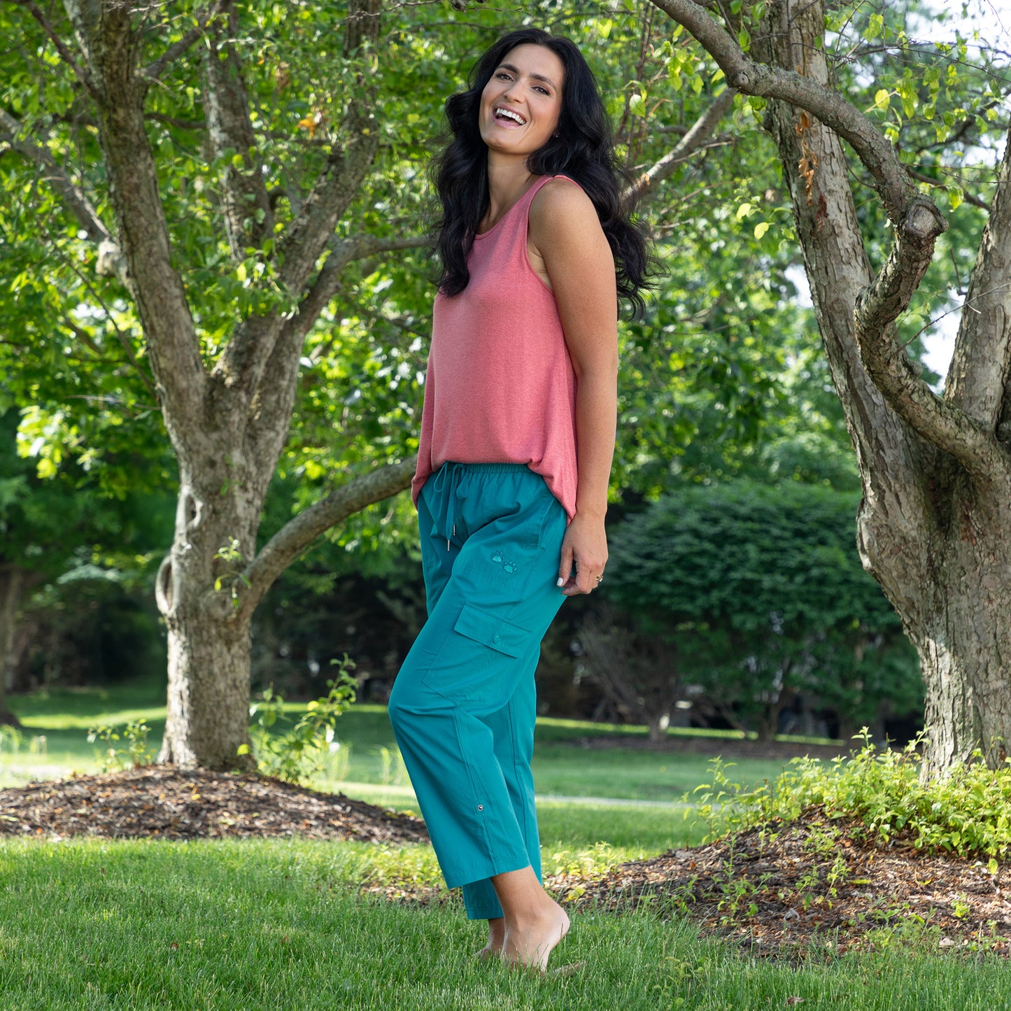 A woman stands barefoot on a grassy lawn in a park, wearing a sleeveless dusty pink top and teal cargo pants.