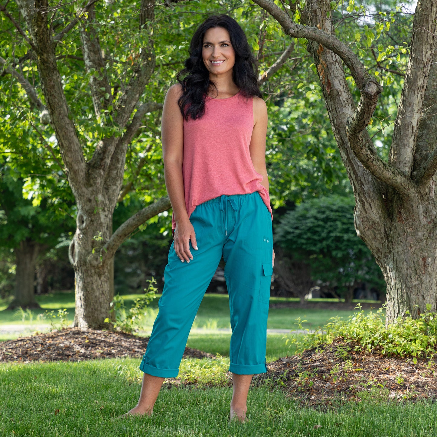 A woman standing outdoors in a park wearing a sleeveless coral pink top and teal cropped cargo pants, with trees and green grass in the background.