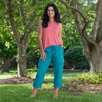 A woman standing outdoors in a park wearing a sleeveless coral pink top and teal cropped cargo pants, with trees and green grass in the background.
