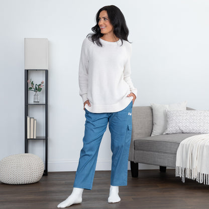 A woman stands in a bright living room wearing a white knit sweater, blue scrub-style pants, and white socks. The room includes a gray sofa, pillows, a floor lamp, and a small shelving unit.