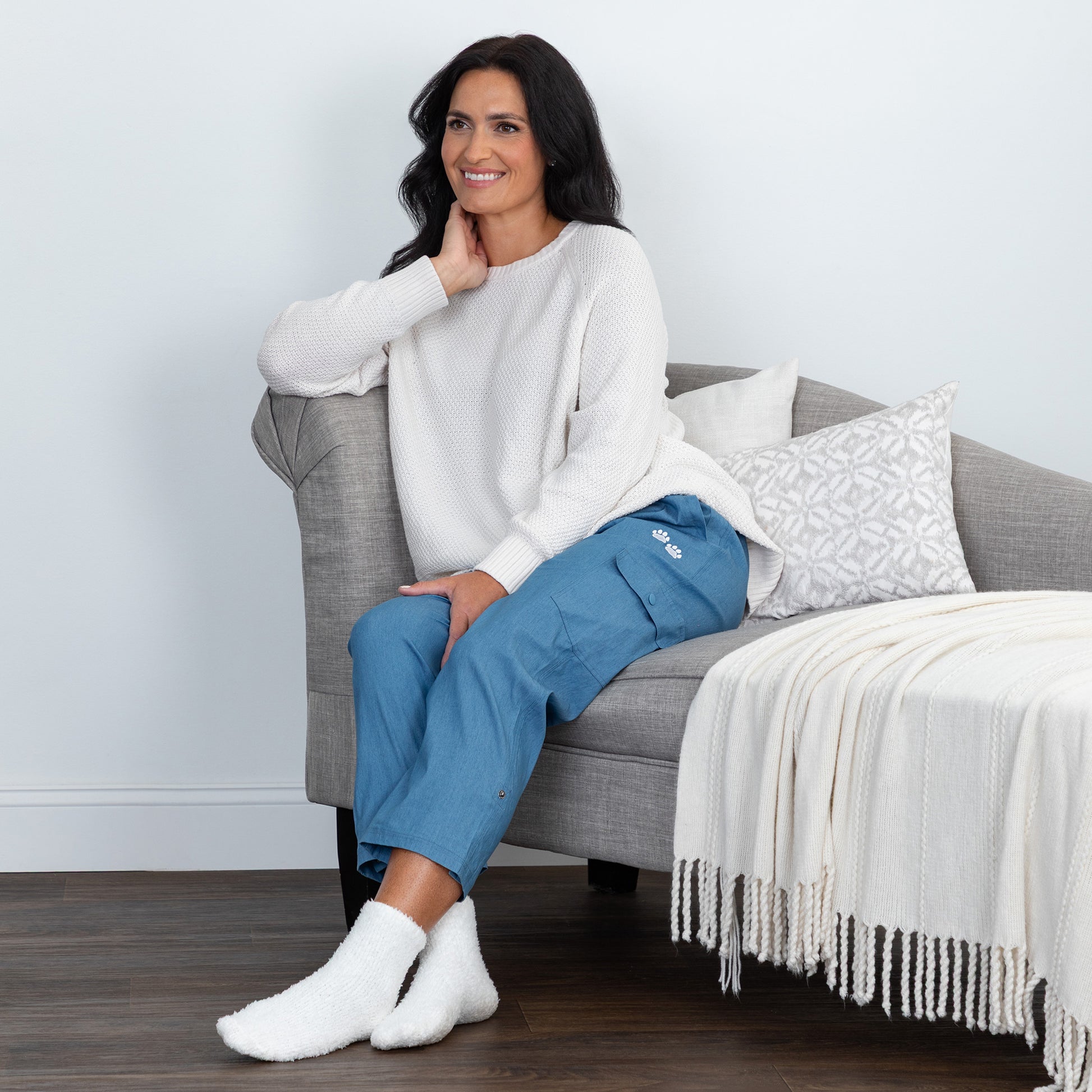 A woman sits on a gray armchair in a living room setting, wearing a white knit sweater, blue cropped pants, and white socks. A white throw blanket with fringe is draped over the chair.