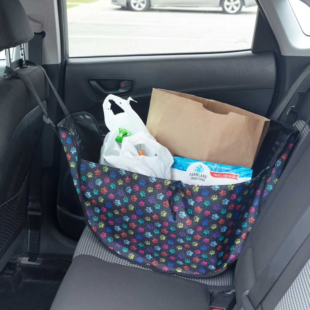 A floral car seat organizer bag filled with grocery items, including a paper bag, plastic bags, and a box of Farmers Supply toilet paper, sitting on a car's back seat.
