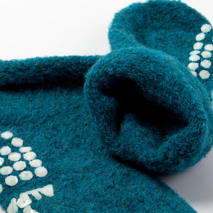 A close-up of a teal blue fuzzy knit textile slipper sock featuring white textured accents on a white background.