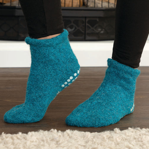 A person wearing plush ankle socks in a bright teal-blue color, standing on a dark wood floor with a white shag rug in the foreground.