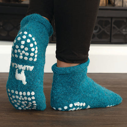 A person wearing teal fuzzy slipper socks with white grip dots and a white paw print on the sole, standing on a dark wood floor.