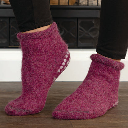 A pair of fuzzy purple felt slipper socks worn on feet, shown from the side against a dark fireplace and wood floor background.