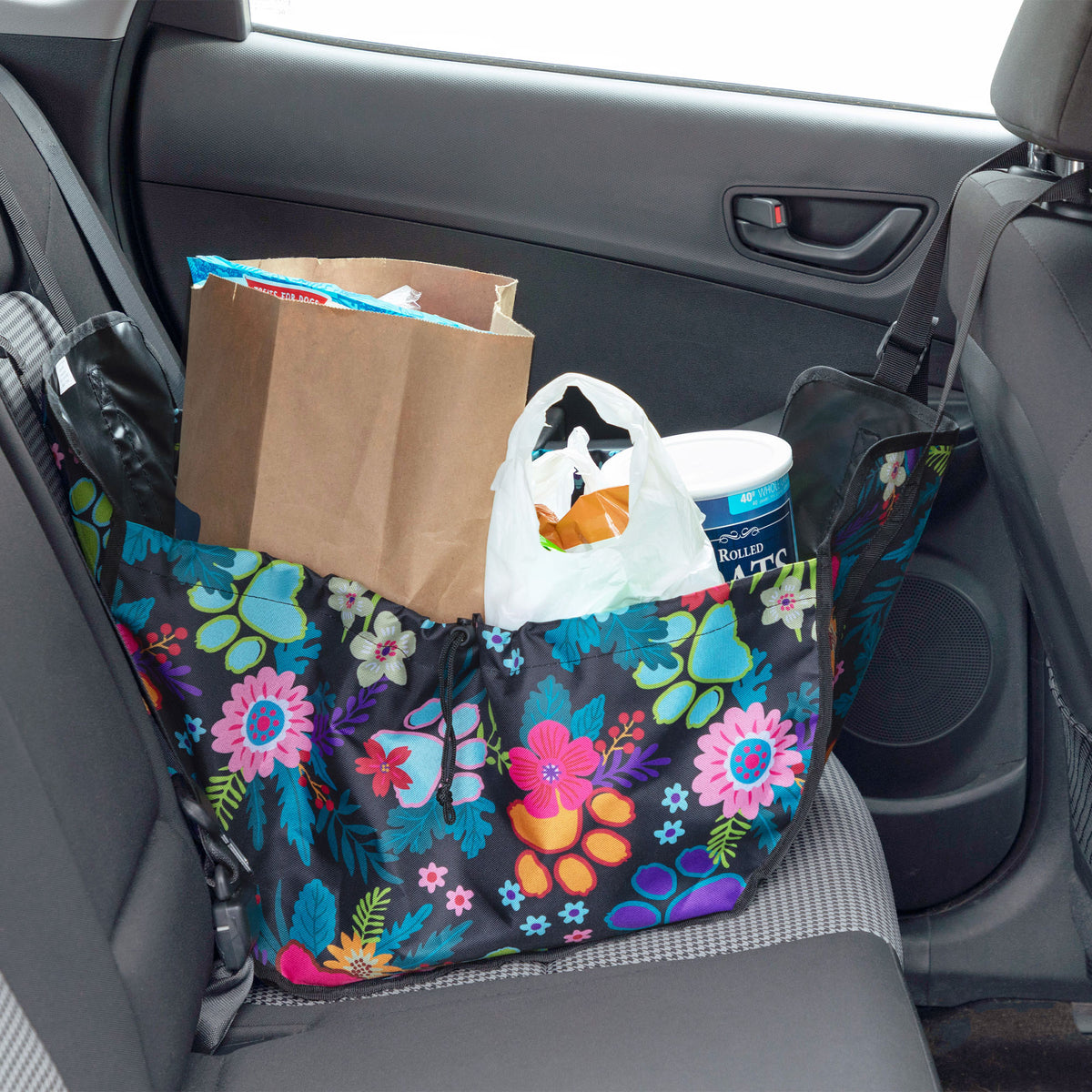 A floral car seat organizer bag filled with grocery items, including a brown paper bag, a white plastic bag, sitting on a gray car seat next to the car door.