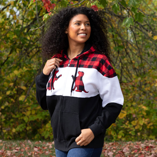 A woman wearing a plaid zip hoodie with dog silhouette details on the front, with trees and fallen leaves in the background.
