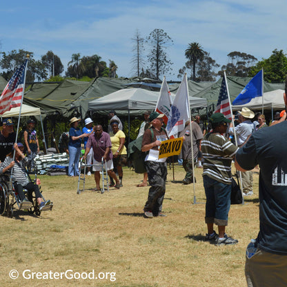 Outdoor event with people and tents, including a person holding a 'Bravo' sign.