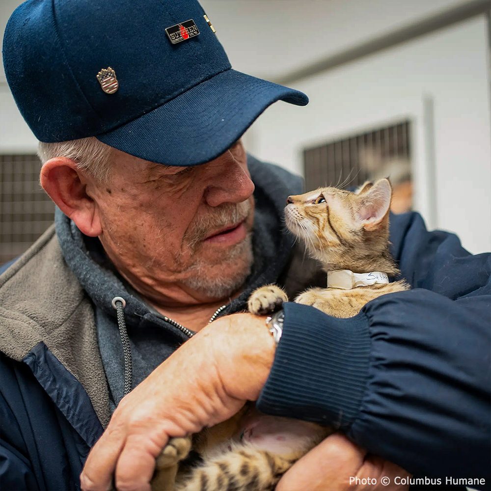 Man holding a small kitten in his arms, wearing a blue cap and coat.