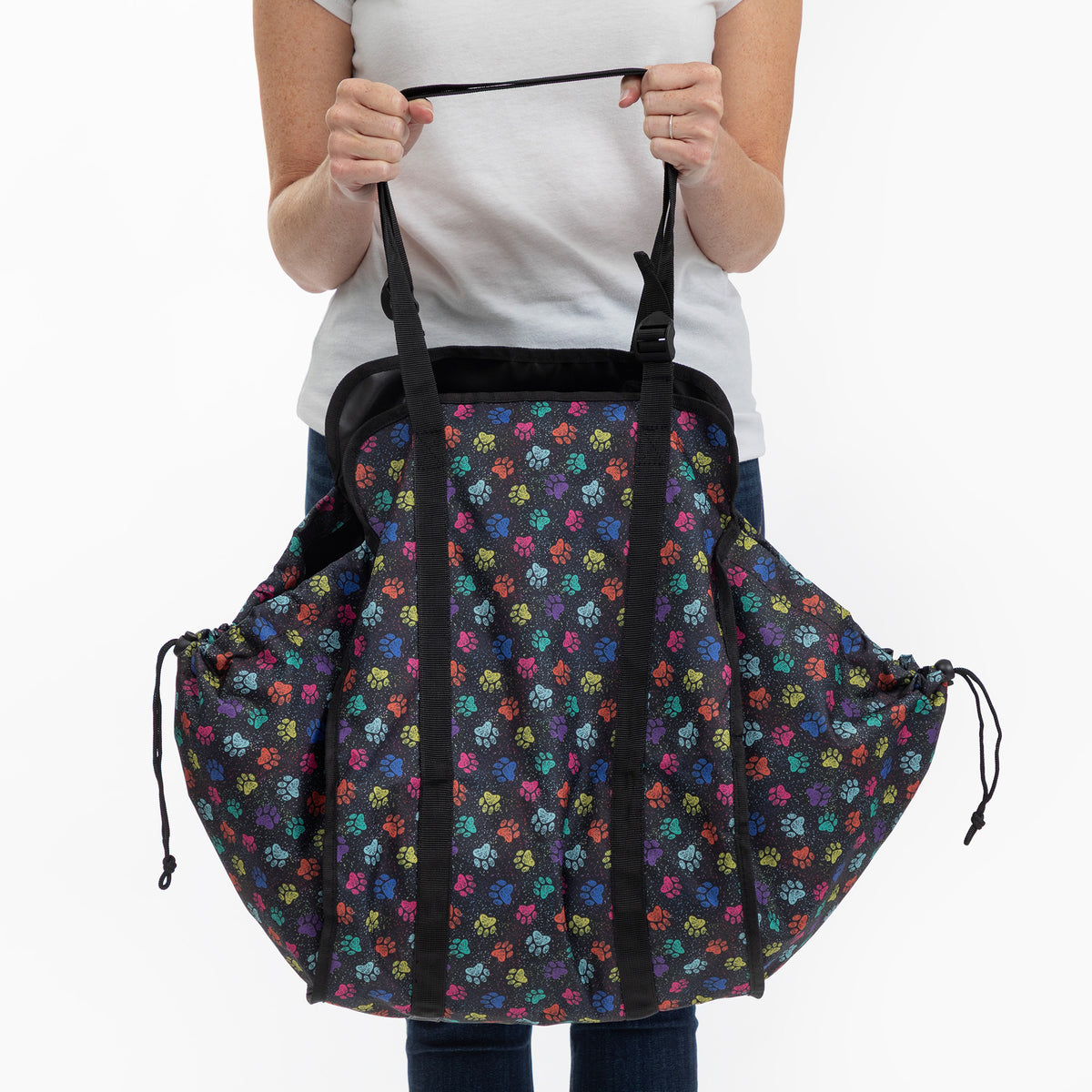 A person holding a large black drawstring collapsible bag with a colorful repeating paw print pattern, shown against a plain white background.