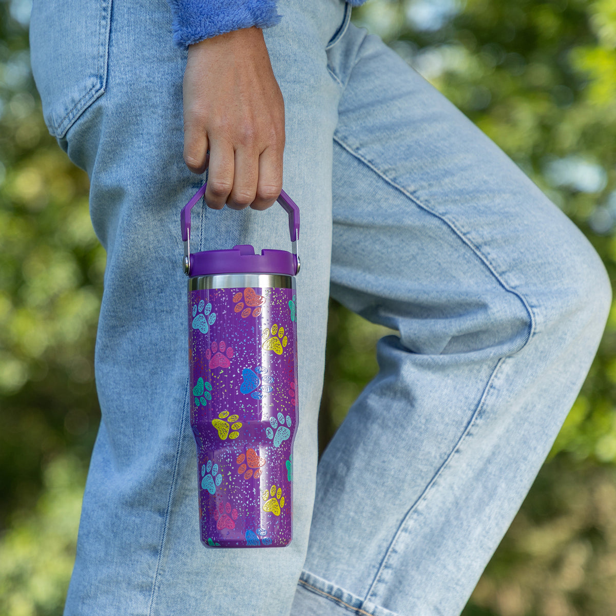 A person wearing light blue jeans holds a purple insulated tumbler with a handle and a colorful paw-print pattern against a blurred outdoor background.