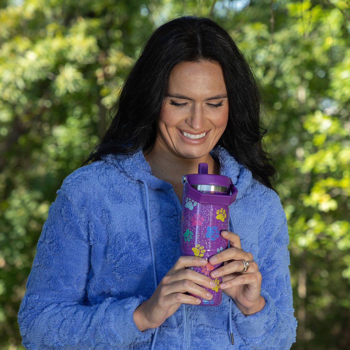 A woman with long black hair wearing a fuzzy blue hoodie is smiling while holding a purple insulated tumbler decorated with colorful paw prints, set against a blurred green outdoor background.