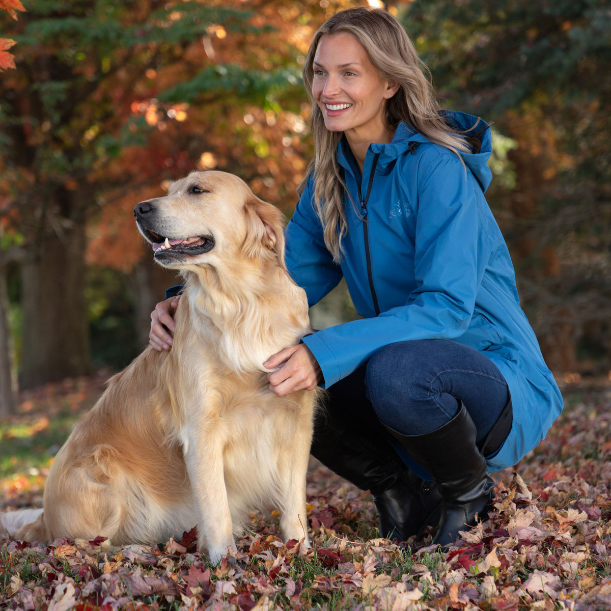 A woman crouches outdoors on a leaf-covered ground next to a golden retriever dog, wearing a bright blue hooded jacket, dark jeans, and black boots. Trees with autumn foliage are blurred in the background.