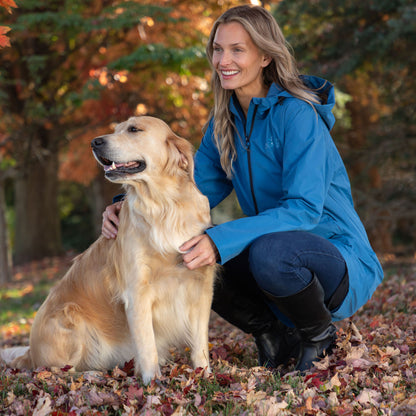 A woman crouches outdoors on a leaf-covered ground next to a golden retriever dog, wearing a bright blue hooded jacket, dark jeans, and black boots. Trees with autumn foliage are blurred in the background.