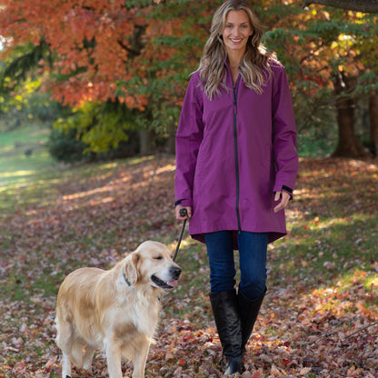 A woman wearing a purple waterproof raincoat walks with a golden retriever dog on a leaf-covered path in an autumn park.