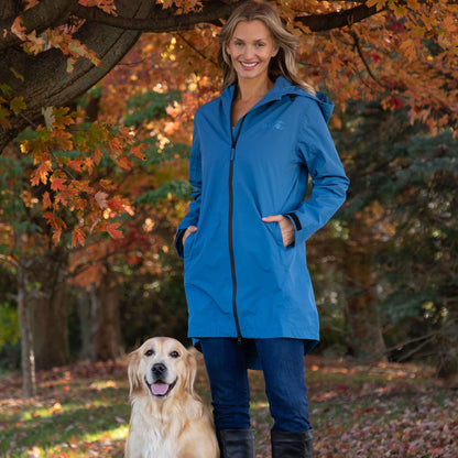 A woman wearing a bright blue hooded raincoat stands outdoors under an autumn tree with a golden retriever dog at her side. The raincoat has a front zipper, large front pockets, and a small tonal logo on the left chest.