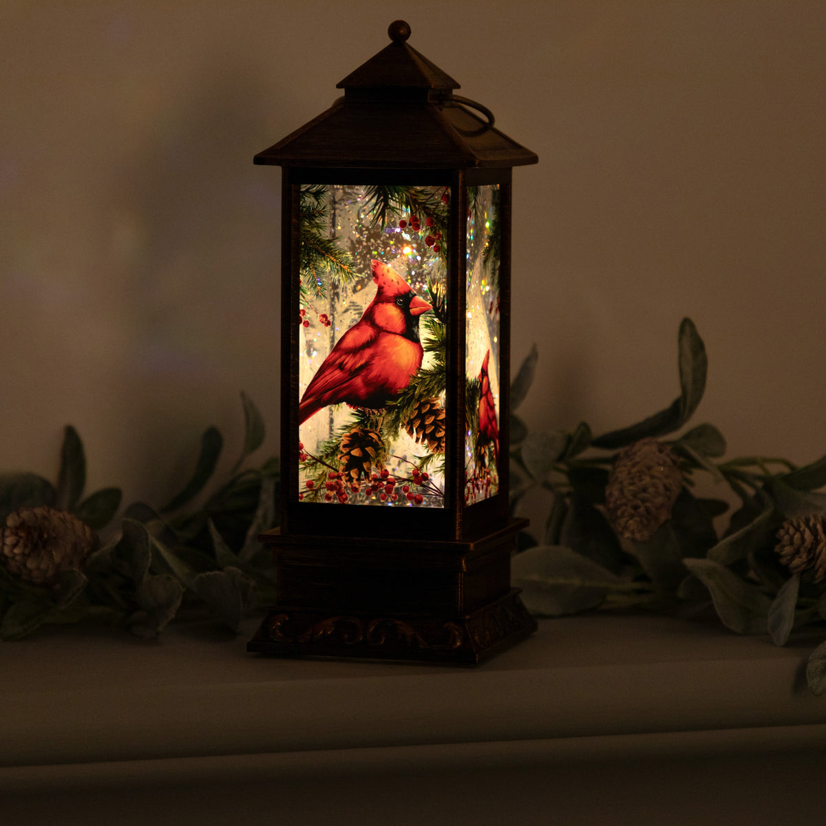 A dark metal lantern with a lit Christmas scene featuring a red cardinal perched on a pine tree, displayed on a shelf with holiday greenery.