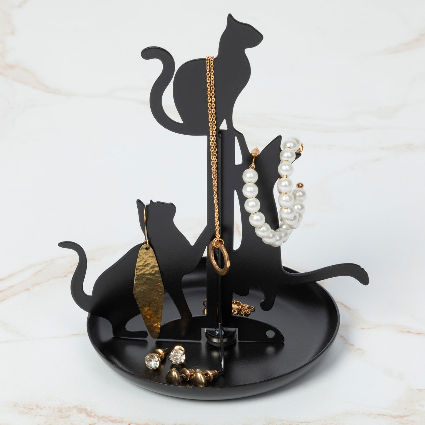A black cat-shaped jewelry organizer stand with silhouettes of three cats, displayed on a light marble surface. The stand holds a gold chain necklace, a white pearl bracelet, a gold earring, and small clear rhinestone earrings.