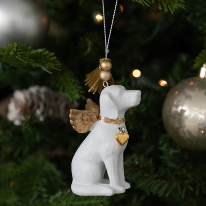 A white dog-shaped Christmas ornament with gold angel wings, a gold collar, and a gold heart-shaped tag, hanging on a decorated Christmas tree.