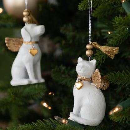 Two white angel-shaped dog and cat Christmas ornaments hanging on a green Christmas tree with warm string lights. Each ornament has gold-colored wings, a collar, a small heart-shaped charm, and a tassel on a string.