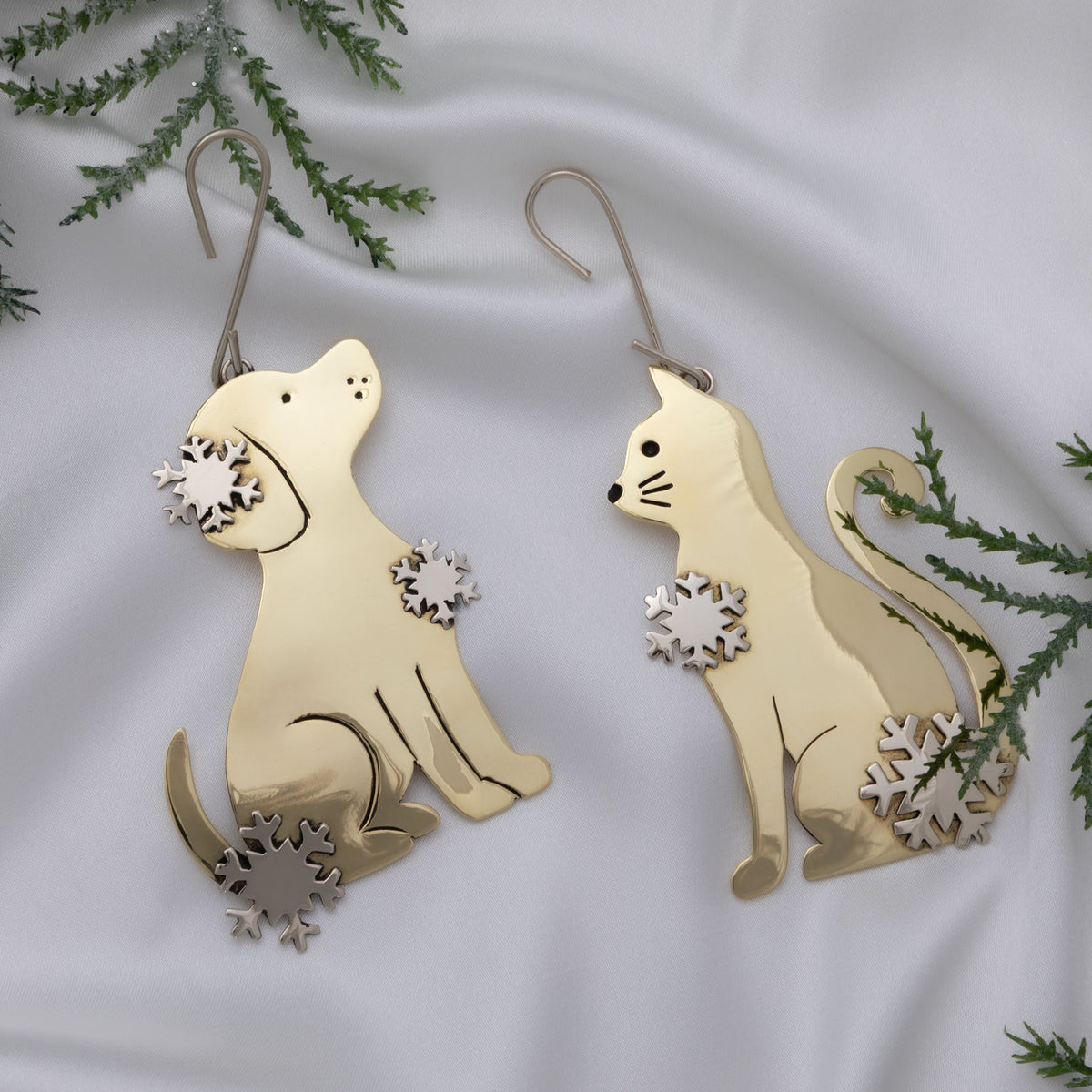 A pair of gold-toned metal ornaments shaped like a sitting dog and a sitting cat, each with small snowflake cutout details, displayed on white fabric with green pine branches around them.