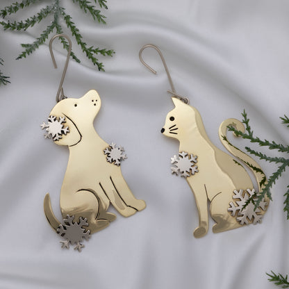 A pair of gold-toned metal ornaments shaped like a sitting dog and a sitting cat, each with small snowflake cutout details, displayed on white fabric with green pine branches around them.