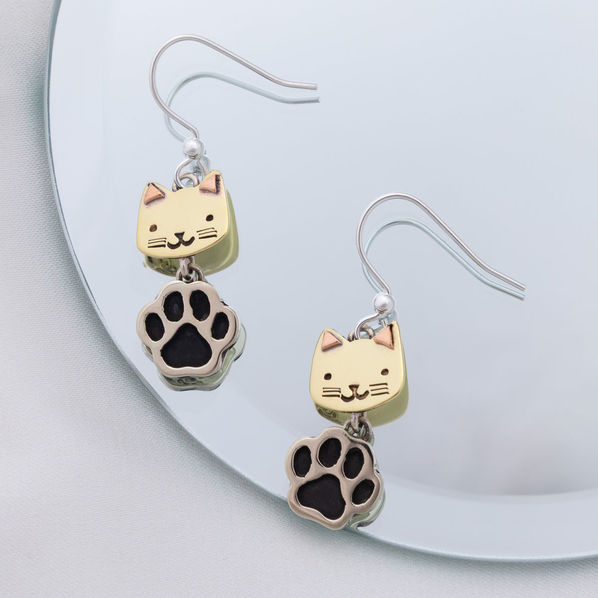 A pair of dangling earrings with small brass cat head charms and black paw print charms, hanging on silver fishhook wires against a pale blue reflective surface.