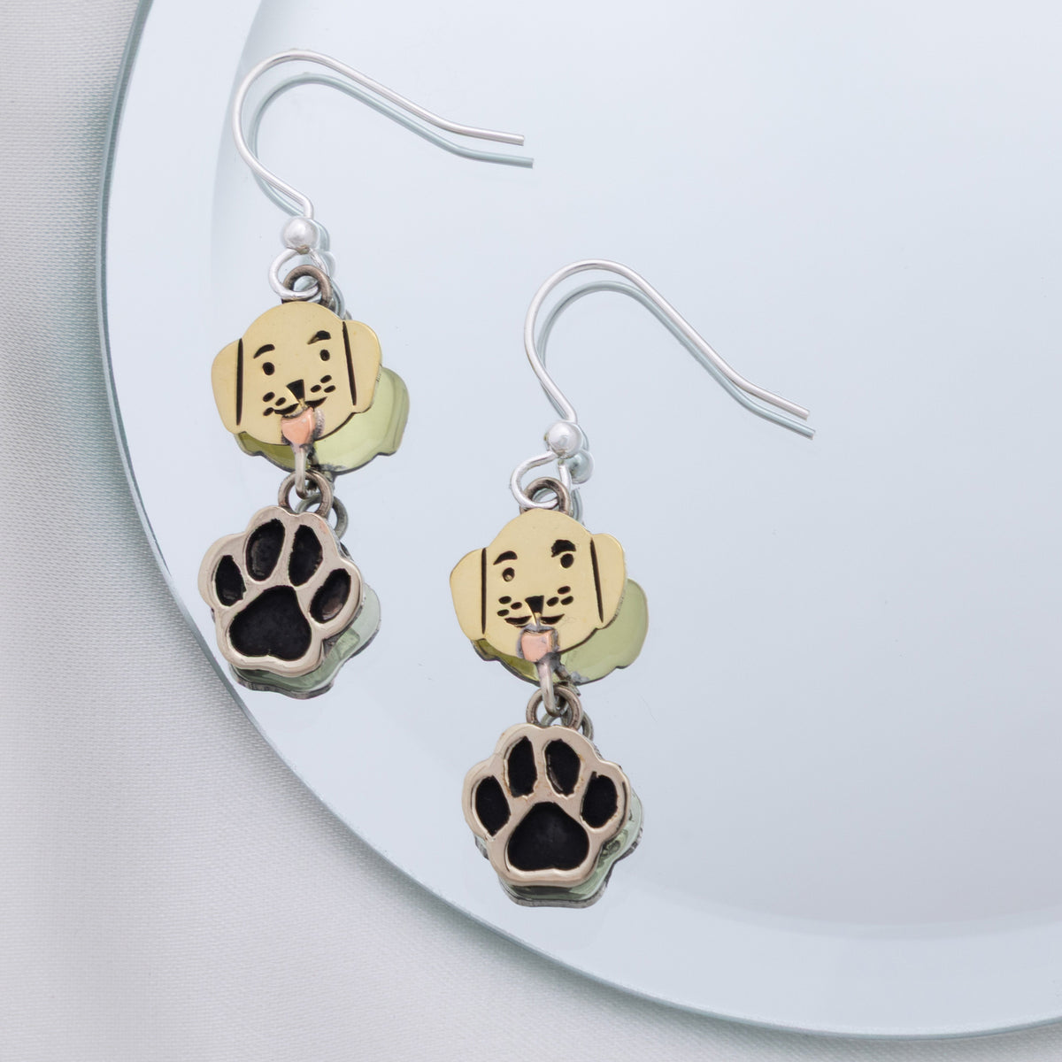 A pair of dangling dog-themed earrings resting on a mirrored surface. Each earring has a silver fishhook wire, a small brass charm shaped like a dog's head with black facial details, and a dangling paw print charm in silver with a black center.