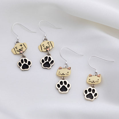Four silver dangle earrings displayed on a soft white fabric background. The set includes two pairs of dog-themed earrings with beige dog head charms and black paw prints, and two pairs of cat-themed earrings with beige cat head charms and black paw prints.