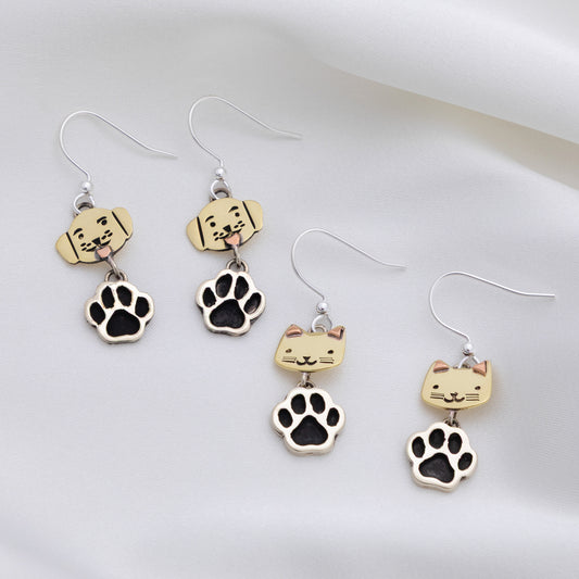 Four silver dangle earrings displayed on a soft white fabric background. The set includes two pairs of dog-themed earrings with beige dog head charms and black paw prints, and two pairs of cat-themed earrings with beige cat head charms and black paw prints.