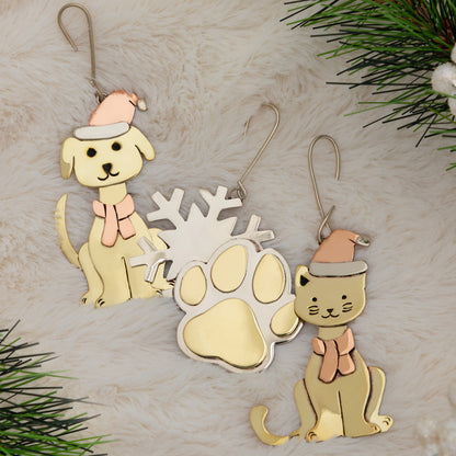 Three Christmas ornaments shaped like a dog, a cat, and a paw print, decorated with pastel colors and hanging hooks, displayed on a white faux fur background with pine branches in the corners.