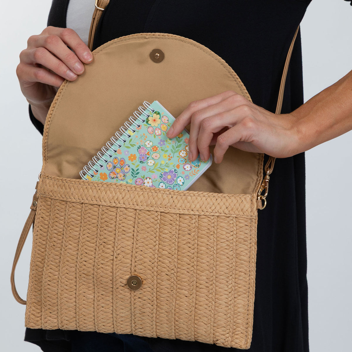 A tan woven crossbody bag with a front flap pocket and a curved top opening, shown being worn by a person. The person's hands are visible opening the bag and placing a small floral notebook inside.