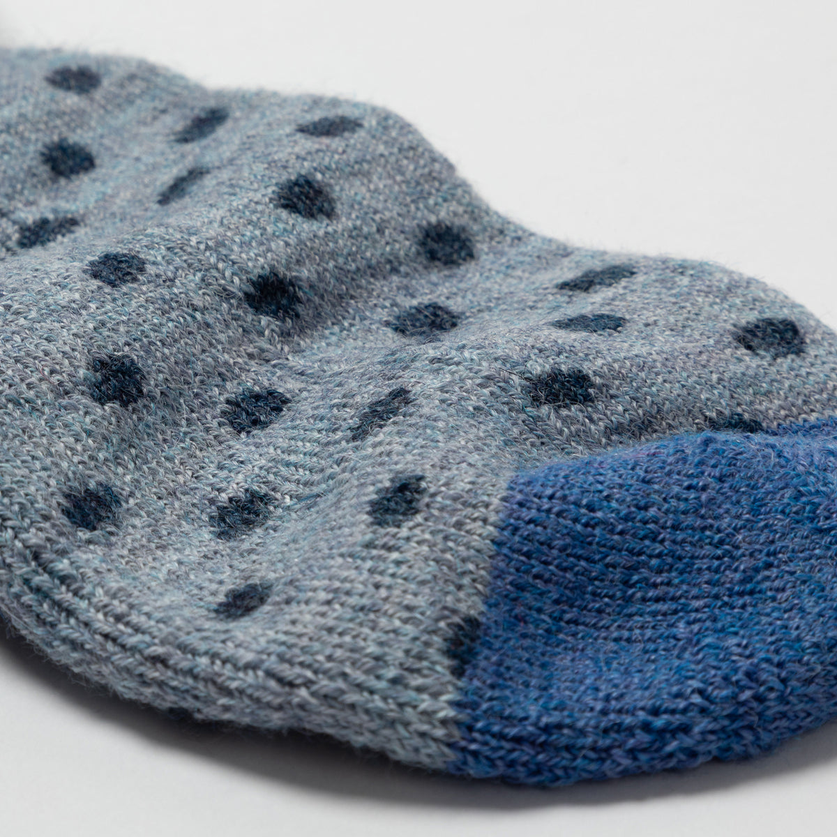 A close-up of a knit sock in heathered light blue-gray with dark blue polka dots and a solid medium blue toe section, photographed on a light neutral background.