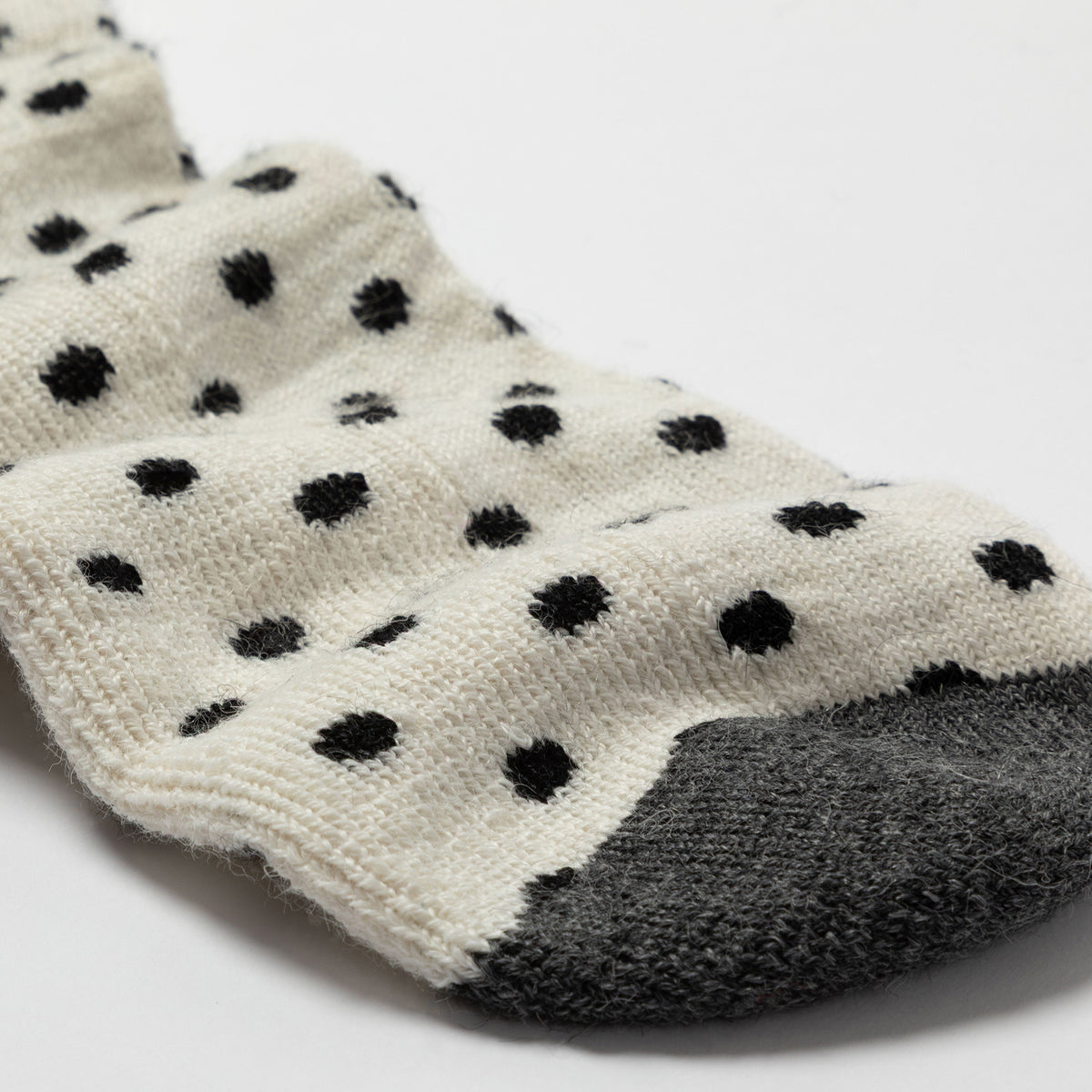 A close-up of a knit sock with black polka dots on an off-white background and a dark gray toe section, photographed on a clean white surface.