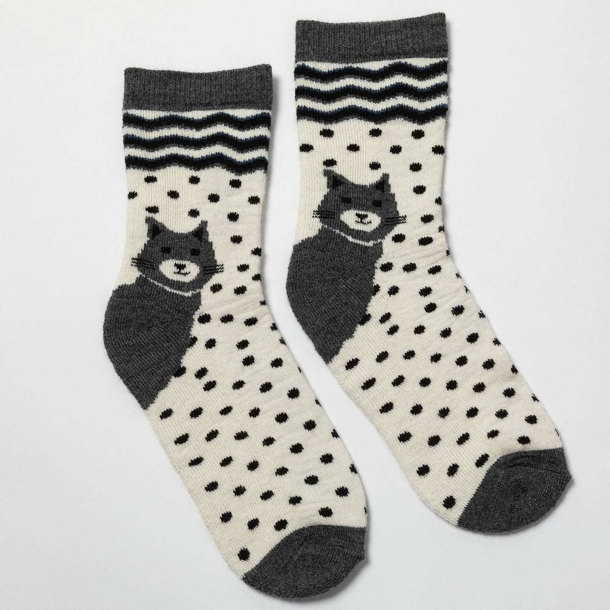 A pair of crew socks with a cream base, black polka dots, dark gray cat illustrations, and black chevron stripes near the ribbed cuffs, shown on a light gray background.