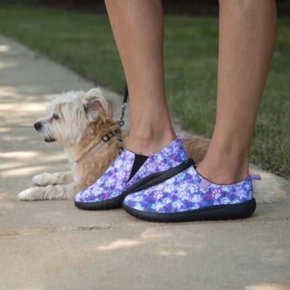 A person wearing slip-on shoes with a purple and blue floral pattern walks outdoors with a small fluffy dog on a concrete path.