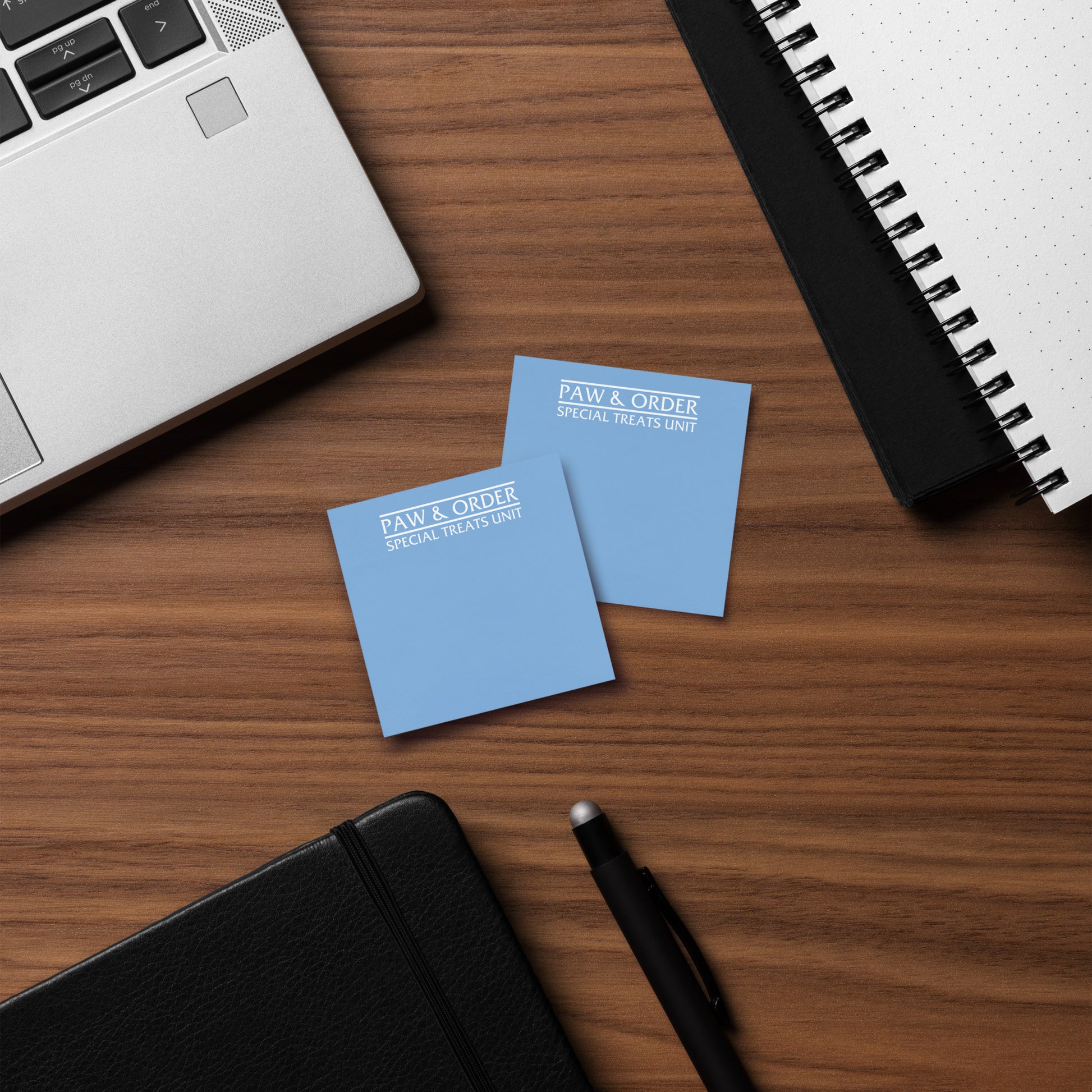 Two sticky note pads with 'Paw & Order Special Treats Unit' text on a wooden surface with a laptop and notebook.