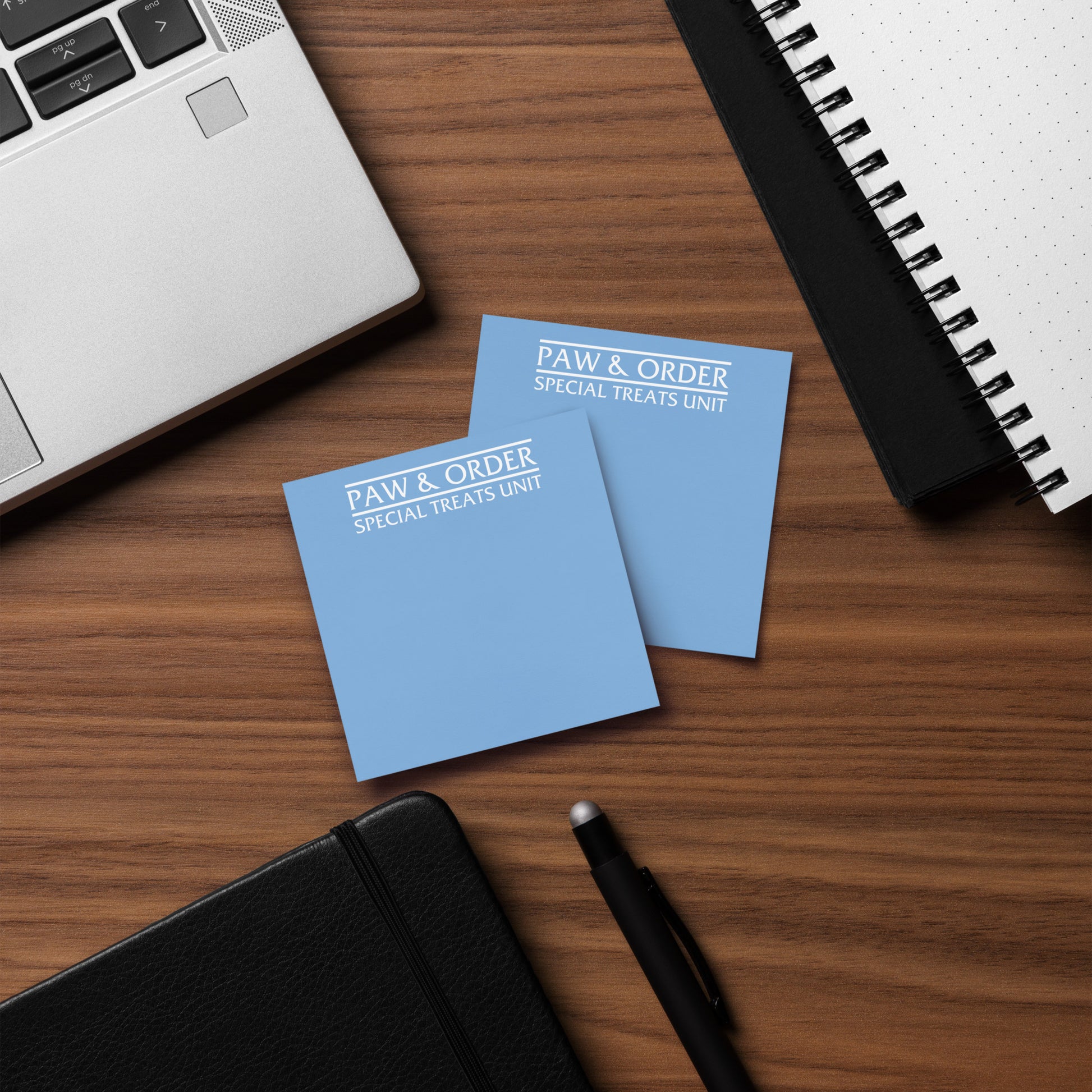 Two sticky note pads with 'Paw & Order Special Treats Unit' text on a wooden surface with a laptop and notebook.