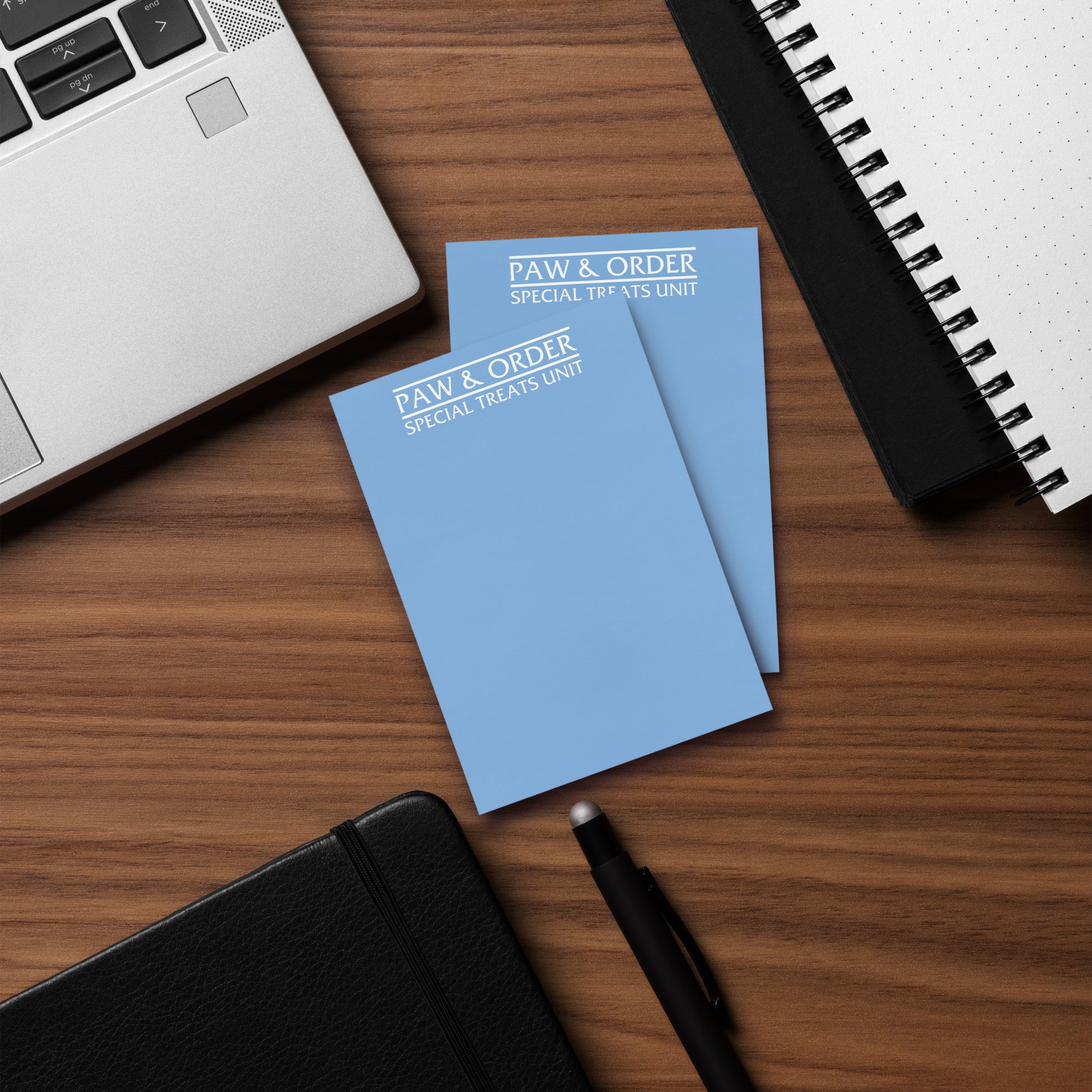 Two longer blue notepads with 'PAW & ORDER' text on a wooden desk with a laptop and pen.