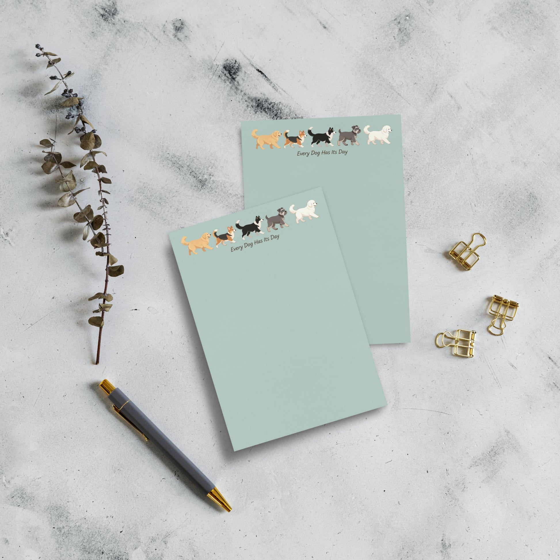 Two 4x6 pastel green notepads with a row of dog illustrations and the text 'Every dog has its day' on the top page, arranged on a light marble surface with a pen, dried stems, and gold binder clips.
