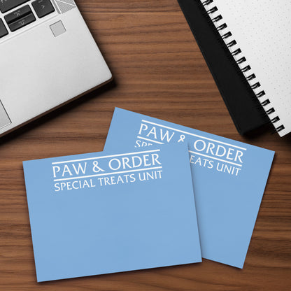 Closeup of Two blue cards with 'Paw & Order Special Treats Unit' text on a wooden surface with a laptop and notebook.
