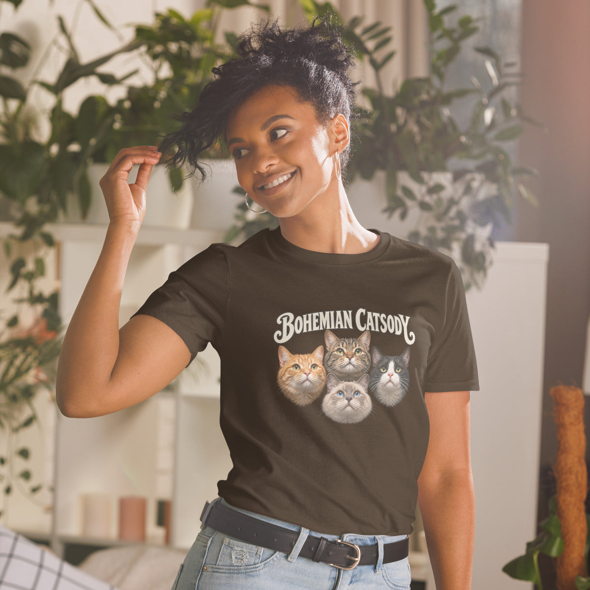 A woman wearing a dark brown graphic t-shirt with the text 'BOHEMIAN CATSODY' and three cat illustrations, paired with light blue jeans in a brightly lit room with indoor plants.