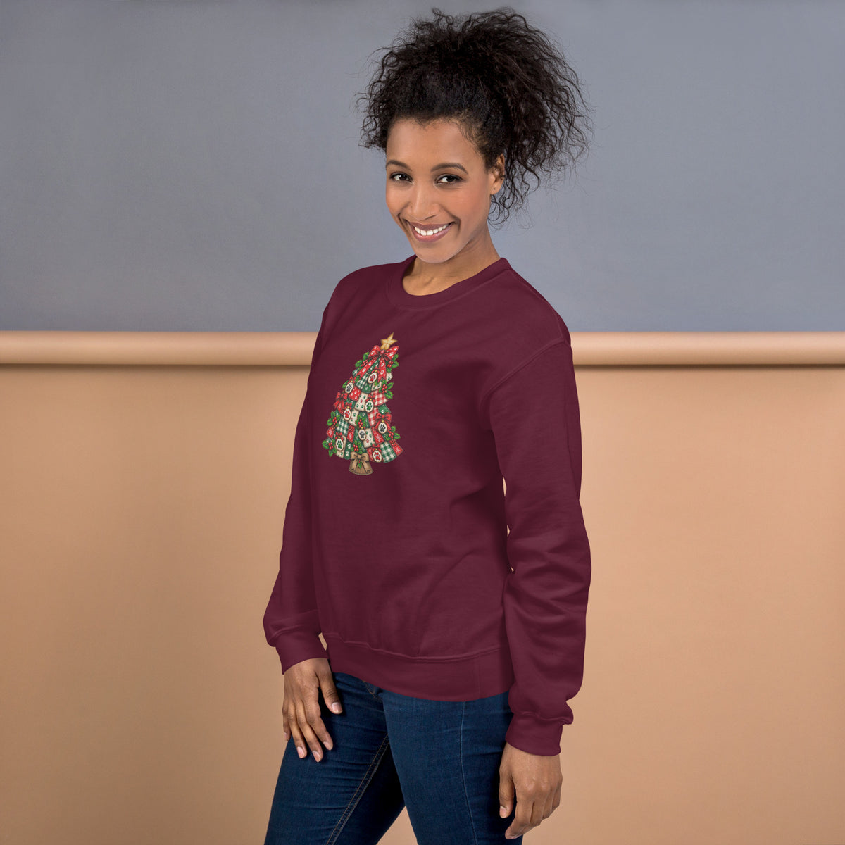 A woman wearing a maroon crewneck sweatshirt with a colorful Christmas tree graphic on the front, paired with blue jeans, standing against a two-tone gray and beige studio background.