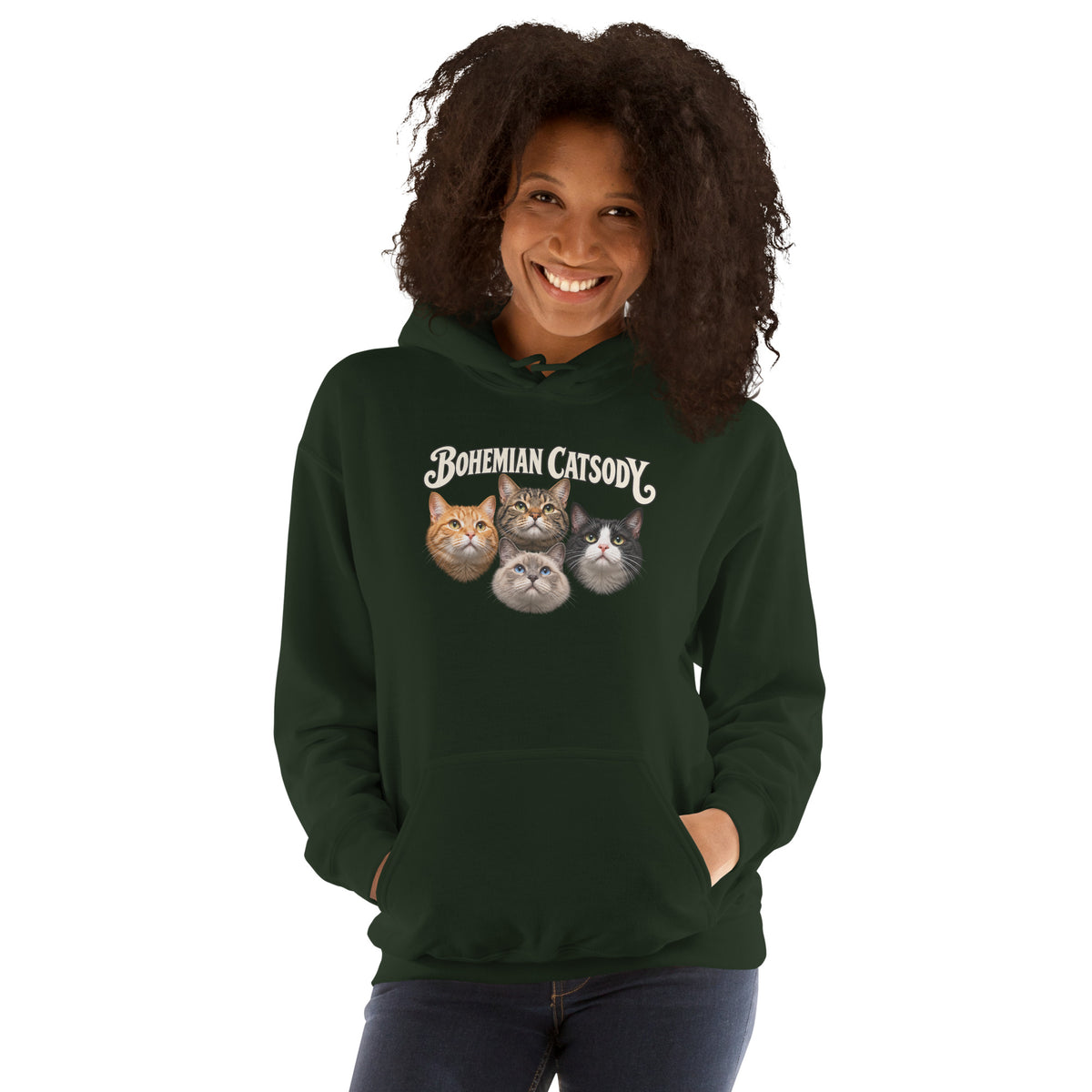 A smiling woman with curly dark hair wears a dark green pullover hoodie featuring a graphic print that reads 'BOHEMIAN CATSODY' above four illustrated cat faces. She is standing against a plain white studio background with her hands in the hoodie pockets.