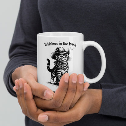 Person holding a white mug with a cat graphic and text 'Whiskers in the Wind' on a plain background