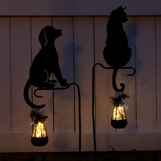 Two solar garden stakes, one in the silhouette of a dog and the other in the silhouette of a cat, each with a lantern that contains solar-powered lights glowing warm light.